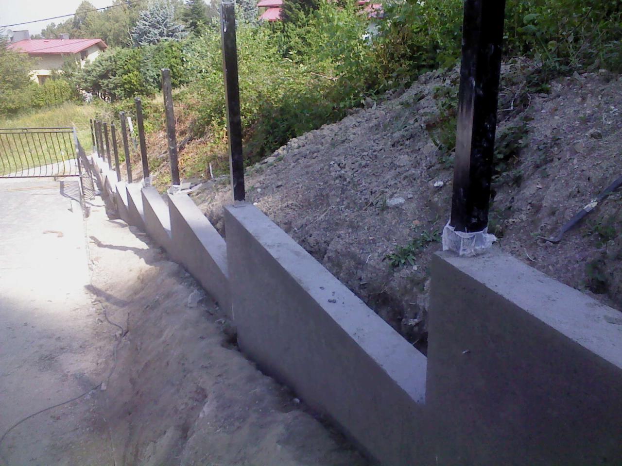 Surowy betonowy murek oporowy z czarnymi słupkami w ziemi, widok z boku, teren nierówny, w tle zieleń i fragment dachu domu.