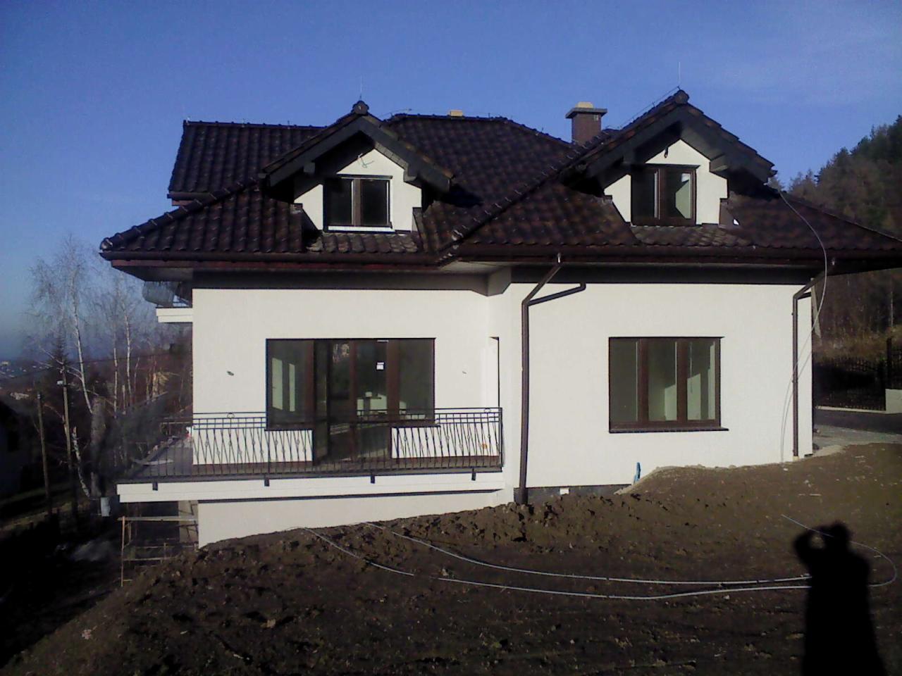 Dom jednorodzinny z balkonem i nową białą elewacją, brązowy dach, widoczne rynny i okna. Na pierwszym planie ziemia.