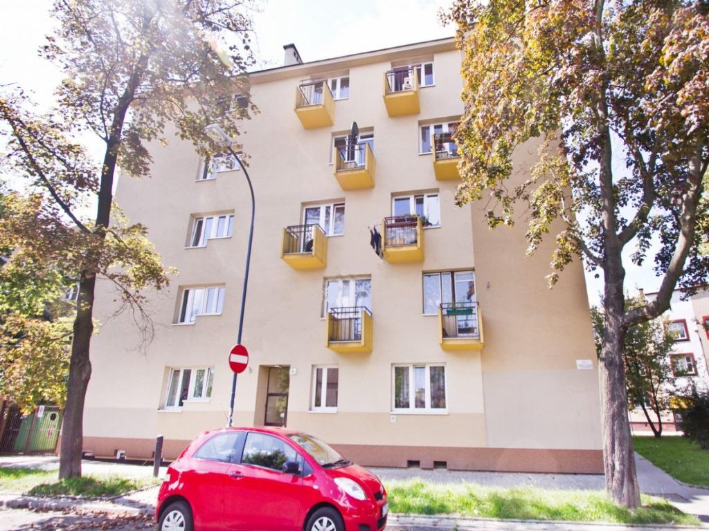 Blok mieszkalny z żółtymi balkonami, stojący obok czerwony samochód osobowy i znak zakazu wjazdu.