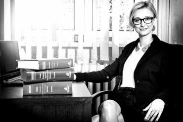 Portret kobiety w okularach siedzącej za biurkiem z książkami prawniczymi i laptopem. Profesjonalny wizerunek prawnika z Wrocławia. Czarno-biała fotografia.