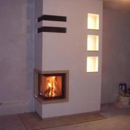 Remax Instalacje elektryczne - Nowoczesny kominek narożny z obudową w jasnym odcieniu, z wbudowanymi półkami i oświetleniem, płomienie widoczne za szybą.