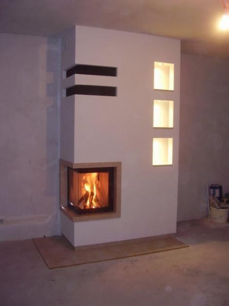 Nowoczesny kominek narożny z obudową w jasnym odcieniu, z wbudowanymi półkami i oświetleniem, płomienie widoczne za szybą.