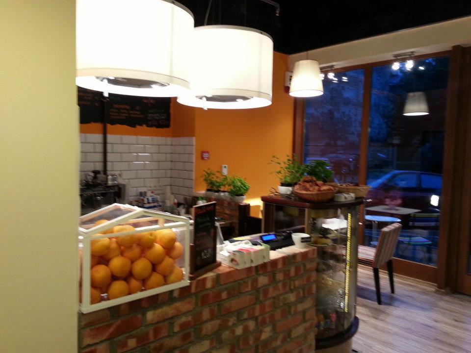 Nowoczesne oświetlenie w kawiarni z pomarańczową ścianą i ceglanym barem. Widoczne wiszące lampy z białymi abażurami i przeszklona witryna z owocami.
