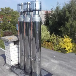 Trzy lśniące, metalowe kominy na płaskim dachu, zamontowane na czarnej, uszczelniającej macie, z ceglanym kominem w tle i zielenią drzew w oddali.