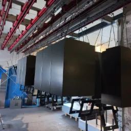 Czarne, prostopadłościenne elementy zawieszone na hakach w hali produkcyjnej, prawdopodobnie po malowaniu proszkowym, widoczne czerwone elementy konstrukcyjne pod sufitem.