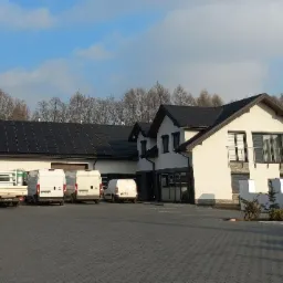 Budynek z panelami słonecznymi na dachu, zaparkowane białe busy dostawcze na brukowanym placu przed budynkiem, błękitne niebo w tle.