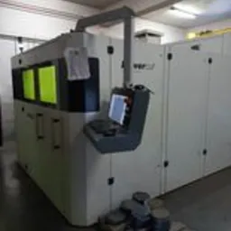 Duża, przemysłowa maszyna do obróbki metali, z panelem sterowania i stosem metalowych elementów na palecie u podstawy. Widoczne żółte światło wewnątrz komory roboczej.