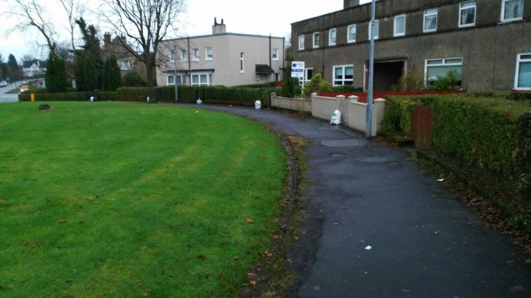 Worki z odzieżą używaną ustawione wzdłuż ścieżki na trawniku przed budynkami mieszkalnymi w pochmurny dzień. Widoczny znak 'For Sale'.