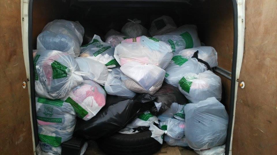 Wnętrze furgonetki wypełnionej workami z odzieżą używaną, częściowo przezroczystymi, z widocznymi ubraniami w środku. Widoczne opony samochodowe.