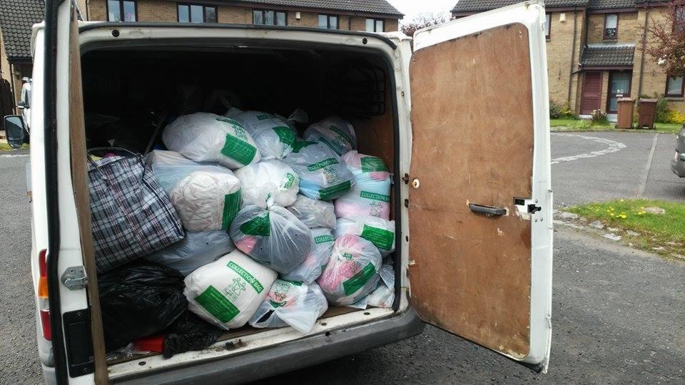 Otwarty bagażnik furgonetki wypełniony workami z odzieżą używaną, gotowymi do transportu. Widoczne budynki w tle.