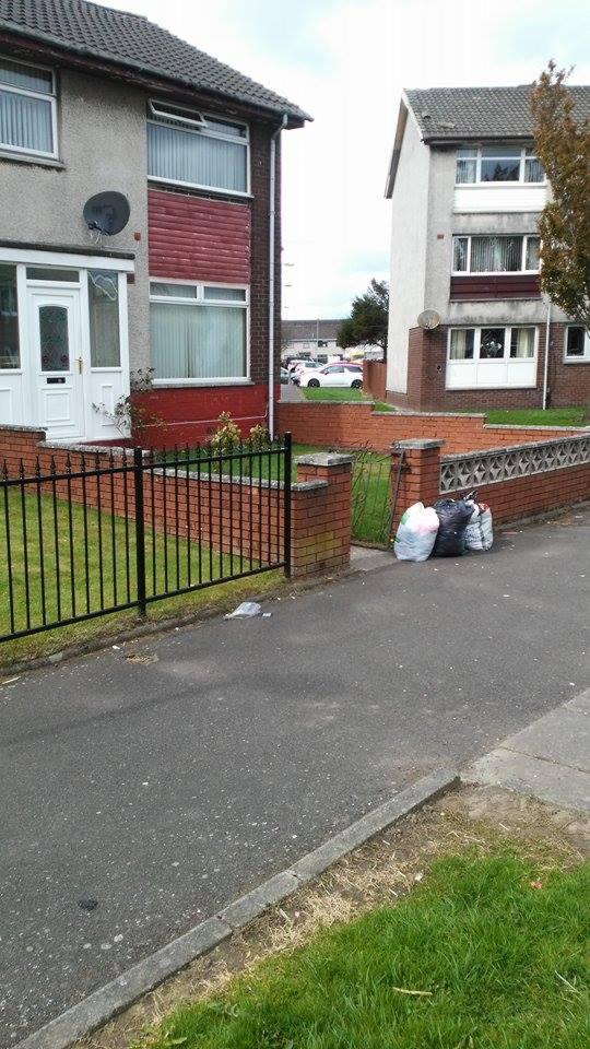 Ulica przed domem z workami, prawdopodobnie z odzieżą, leżącymi przy niskim murku. Widoczne budynki mieszkalne i trawnik.