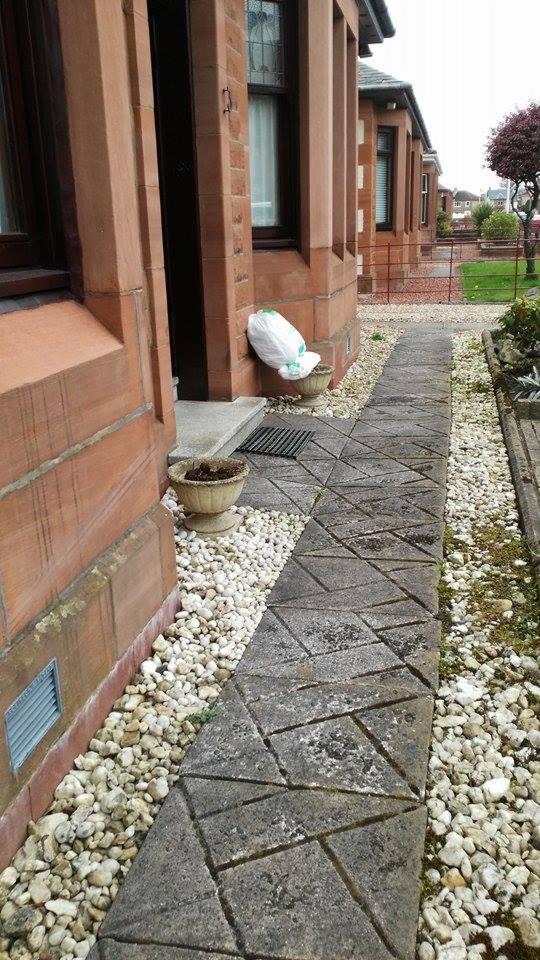 Chodnik z geometrycznych płyt betonowych, obłożony białym żwirem, prowadzący do wejścia domu z czerwonej cegły. Przy drzwiach donica i biały worek.