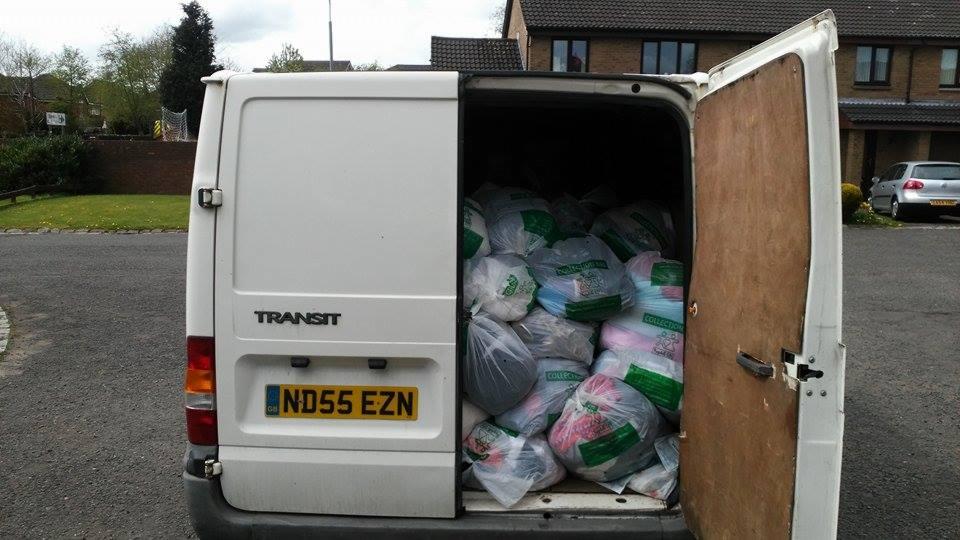 Tył białego furgonetki marki Transit z otwartymi drzwiami, wypełnionego workami z odzieżą używaną, widoczne tablice rejestracyjne ND55 EZN.