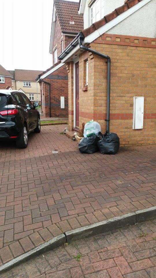 Sterta worków na śmieci przed domem z cegły, obok zaparkowany czarny samochód. Widok na brukowany podjazd i fragment budynku z rynnami.