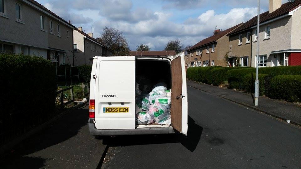 Biały van z otwartymi drzwiami, wypełniony workami z odzieżą używaną, zaparkowany na ulicy w dzielnicy mieszkalnej w słoneczny dzień.
