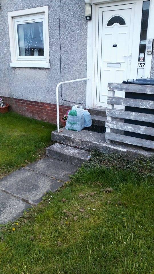 Dwa worki z odzieżą używaną stoją na schodach przed wejściem do domu z numerem 137. Widoczny fragment chodnika i trawnika. Ujęcie z dołu.