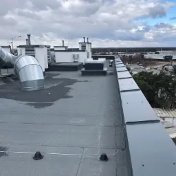 Płaski dach pokryty papą z widocznymi elementami wentylacji, kominami i obróbkami blacharskimi, z widokiem na przemysłowy krajobraz w pochmurny dzień.