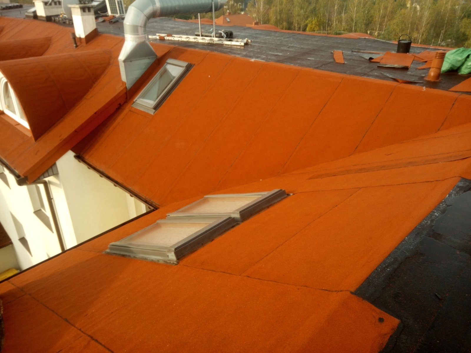 Pomarańczowy dach z papy termozgrzewalnej z oknami dachowymi, widoczne kominy wentylacyjne i fragmenty starego pokrycia, w tle drzewa.
