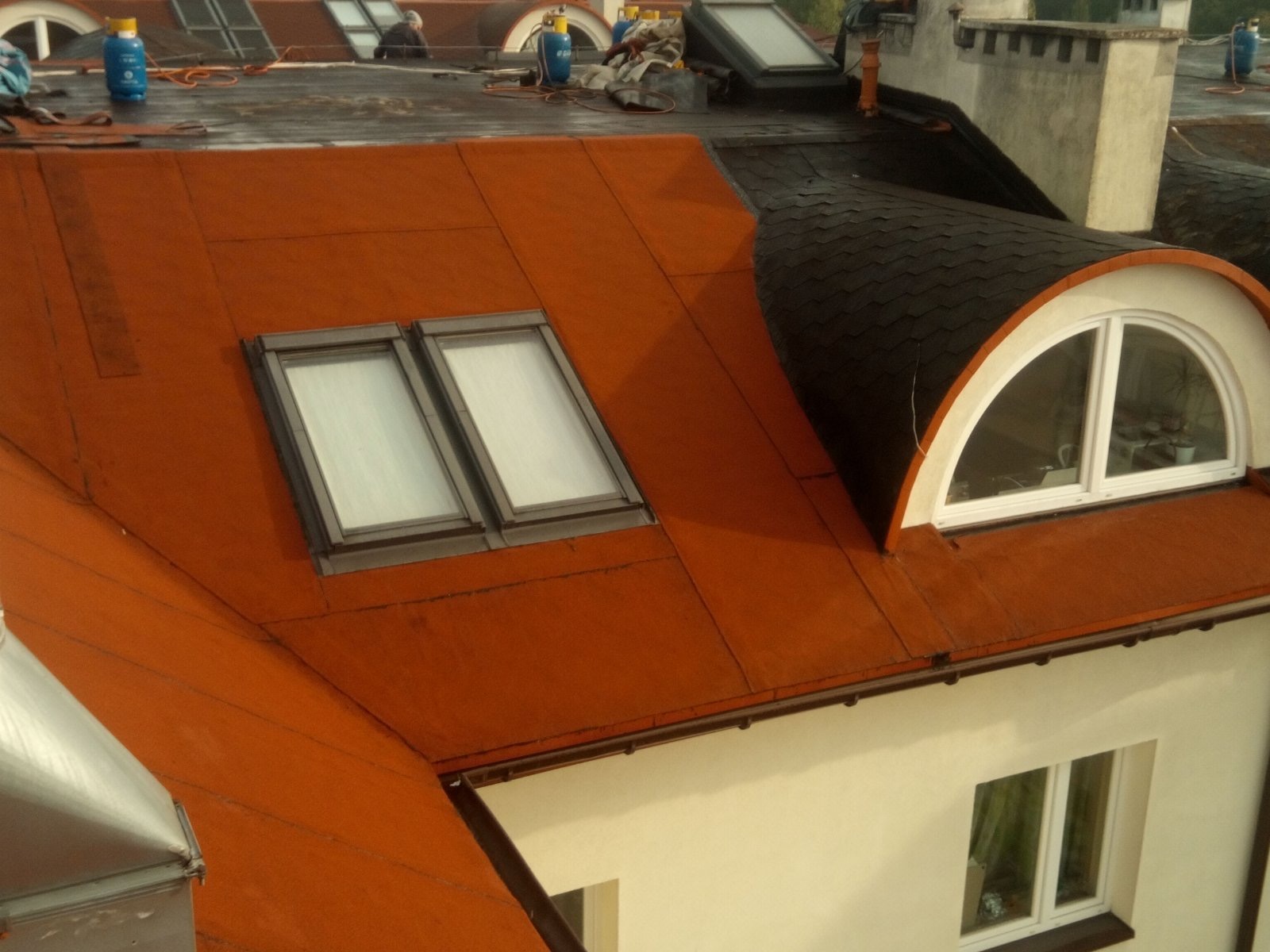 Widok z góry na dach pokryty czerwoną papą z dwoma oknami dachowymi i lukarną krytą czarnym gontem bitumicznym, na dachu widoczne butle gazowe i narzędzia.