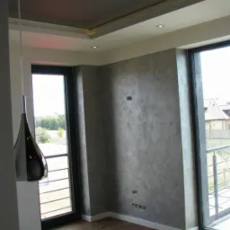 Pokój z wykończonymi ścianami w odcieniach szarości, drewnianą podłogą i nowoczesną lampą wiszącą. Widoczne duże okna z widokiem na zewnątrz i podwieszany sufit z oświetleniem punktowym.