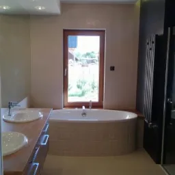 Nowoczesna łazienka z owalną wanną pod oknem, drewnianą szafką pod umywalkami i dekoracyjnym kryształowym żyrandolem sufitowym.