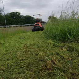 Operator na pomarańczowej kosiarce samojezdnej tnie wysoką trawę przy drodze, widok z tyłu, pochmurne niebo.