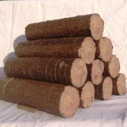 Ułożone w piramidę cylindryczne brykiety z biomasy na tle białego, lekko pogniecionego materiału, z widoczną teksturą i drobnymi pęknięciami na powierzchni brykietów.