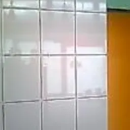 Błyszczące, białe płytki ścienne z ciemną fugą, odbijające światło okna, zestawione z fragmentem pomarańczowej ściany. Minimalistyczny detal wykończenia wnętrza.