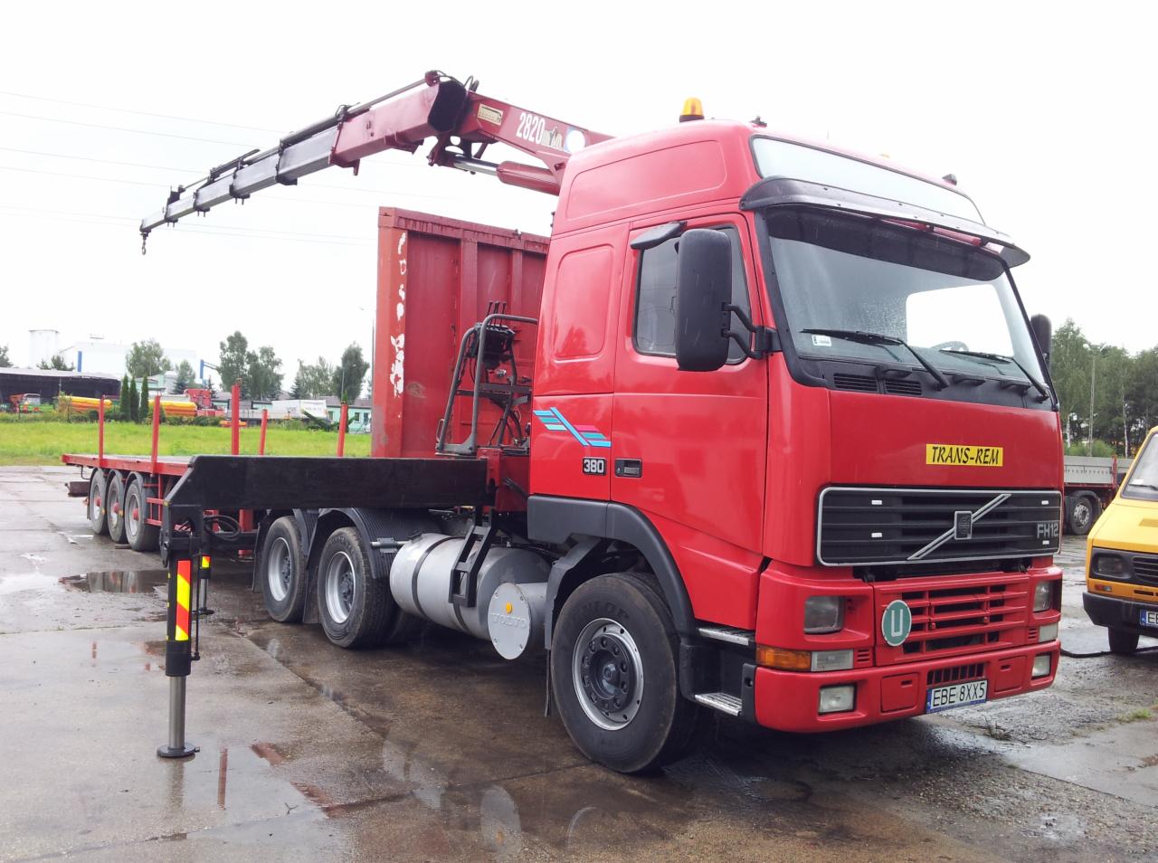 Czerwony samochód ciężarowy Volvo FH12 z dźwigiem i platformą, zaparkowany na mokrym placu, z widocznym logo TRANS-REM na kabinie i wysuniętą podporą stabilizującą.