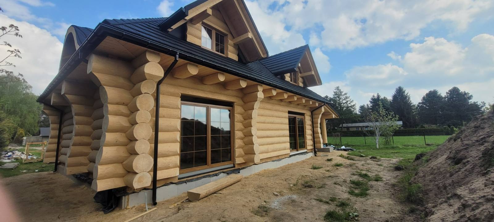 Surowy drewniany dom z bali z czarnym dachem i rynnami, widoczne okna z brązowymi ramami, teren wokół domu w trakcie prac ziemnych.