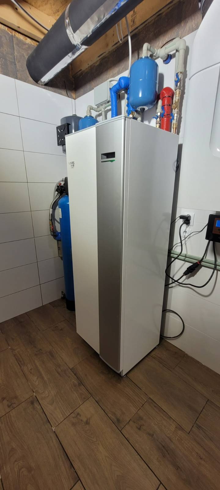 Nowoczesna, biało-srebrna jednostka wewnętrzna z niebieskimi zbiornikami wyrównawczymi i rurami, zainstalowana w pomieszczeniu z drewnopodobną podłogą i białymi płytkami ściennymi.