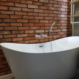 Wolnostojąca biała wanna z chromowaną baterią i prysznicem na tle ściany z cegły i półek w łazience.