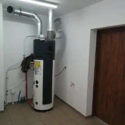 Kompaktowa pompa ciepła z szarymi rurami wentylacyjnymi w pomieszczeniu gospodarczym w Starym Licheniu, widoczna instalacja wodna i odpływ w podłodze.