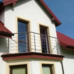 Balkon z balustradą ze stali nierdzewnej, widok z dołu, jasna elewacja budynku z czerwonym dachem, błękitne niebo w tle, nowoczesny design.
