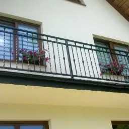 Czarna, metalowa balustrada balkonowa z ozdobnymi elementami, zamontowana na balkonie budynku z białymi ścianami i brązowymi oknami, z kwiatami w doniczkach.