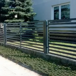 Nowoczesne ogrodzenie metalowe z poziomymi profilami, widok z perspektywy chodnika, z zielonym trawnikiem i domem w tle.