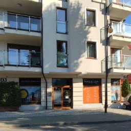 Fasada budynku z biurami finansowymi na parterze, z balkonami na wyższych piętrach, numer 13 nad wejściem, szyldy z hasłami 'Eksperci Finansowi' i 'Finanse bez granic' w oknach.