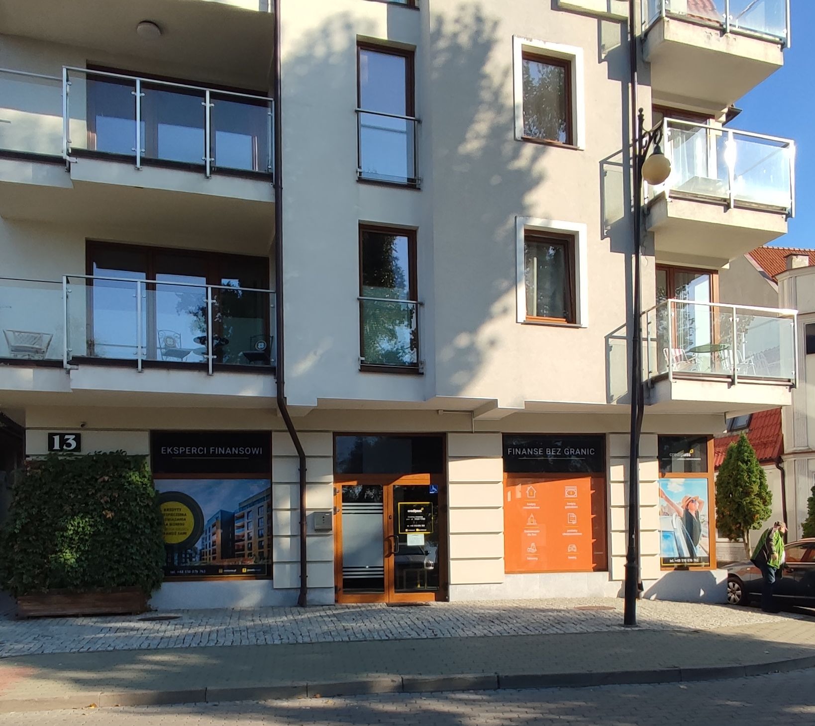 Fasada budynku z biurami finansowymi na parterze, z balkonami na wyższych piętrach, numer 13 nad wejściem, szyldy z hasłami 'Eksperci Finansowi' i 'Finanse bez granic' w oknach.