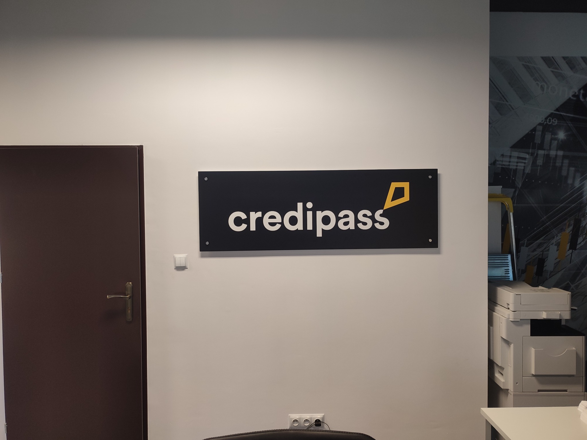 Logo firmy Credipass na czarnym szyldzie, zamontowane na białej ścianie biura, obok brązowych drzwi i z widocznym fragmentem drukarki w tle.