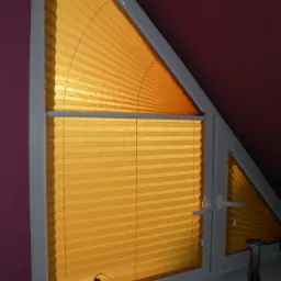 Żółta, plisowana osłona okienna w trójkątnym oknie na poddaszu, z dekoracjami na parapecie.