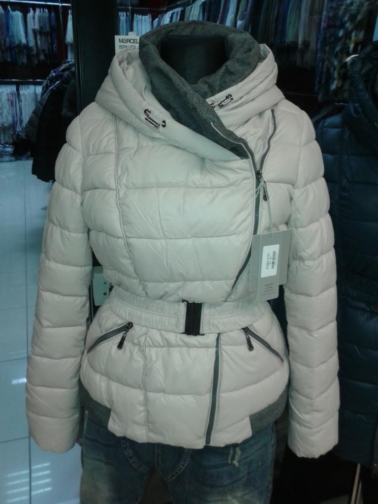 Pudrowa, pikowana kurtka zimowa z asymetrycznym zapięciem i paskiem w talii, prezentowana na manekinie na tle sklepowych wieszaków z odzieżą.