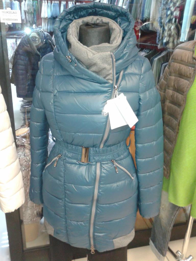 Niebieska, pikowana kurtka damska z kapturem i paskiem, prezentowana na manekinie w sklepie odzieżowym. Widoczne srebrne zamki i metki.