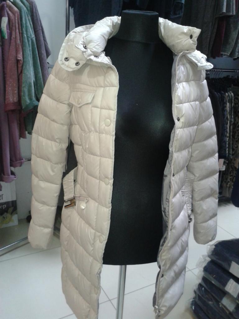 Beżowy, pikowany płaszcz damski z kapturem na czarnym manekinie w sklepie odzieżowym. Widoczne detale: kieszenie, guziki, pasek. W tle wieszaki z ubraniami.