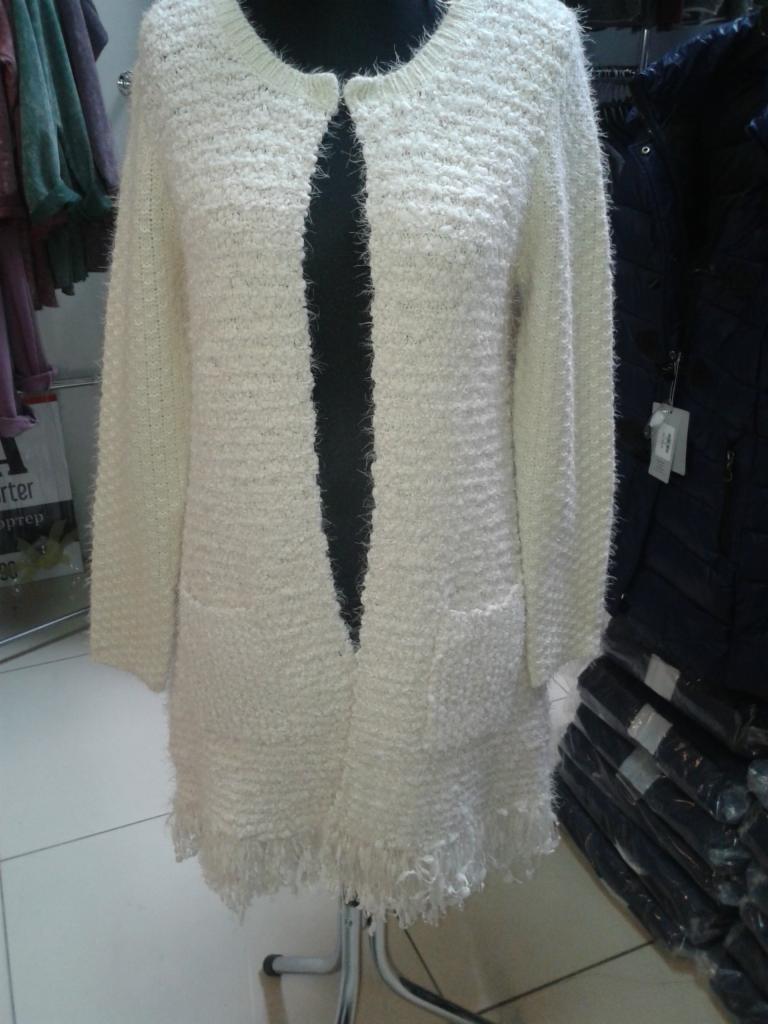 Kremowy, dziergany kardigan z frędzlami na manekinie, widoczne kieszenie i faktura materiału. W tle inne ubrania na wieszakach i złożone w stosy.