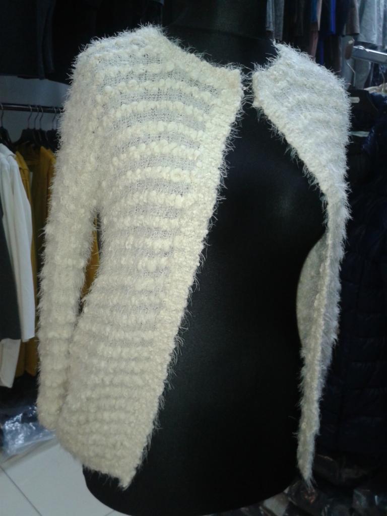 Kremowy, włochaty kardigan bez zapięcia na czarnym manekinie. Widoczne poziome prążki w strukturze dzianiny. W tle wieszaki z innymi ubraniami.