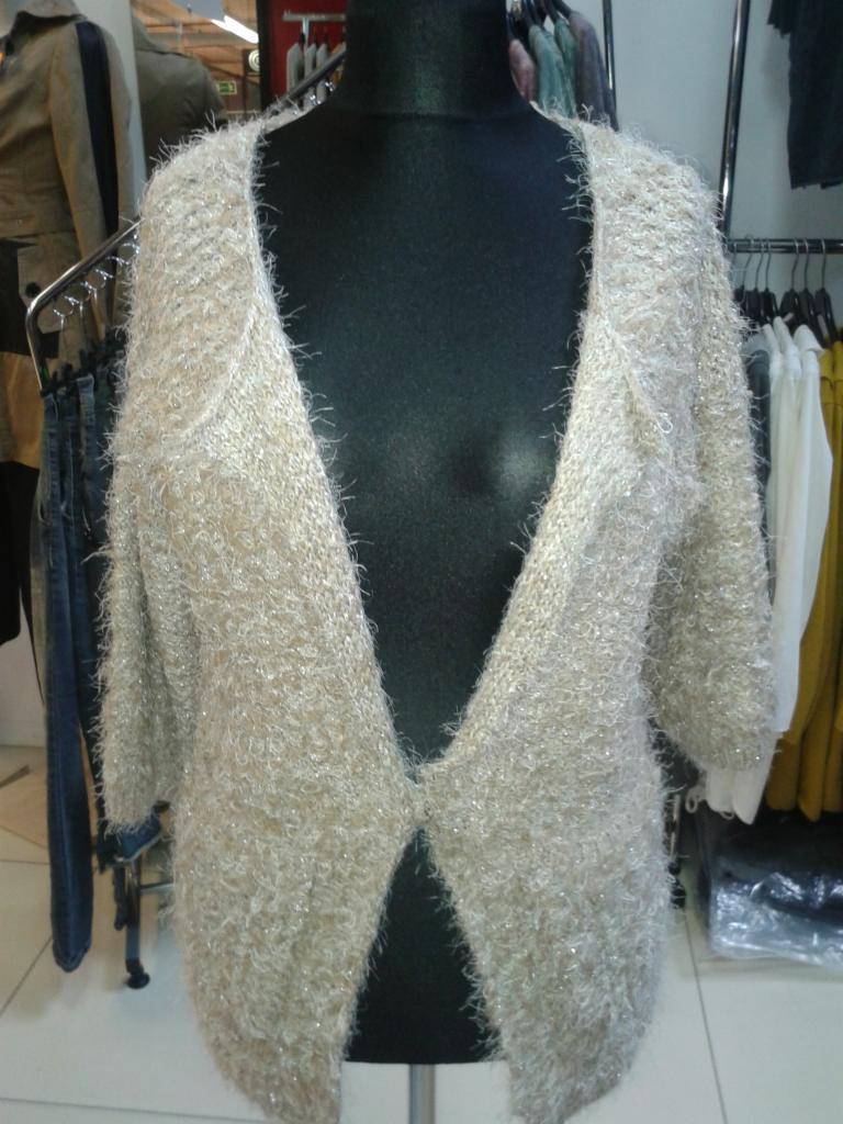 Beżowy, włochaty kardigan z krótkim rękawem na czarnym manekinie, w tle wieszaki z odzieżą. Ujęcie z bliska, prezentujące fakturę materiału.