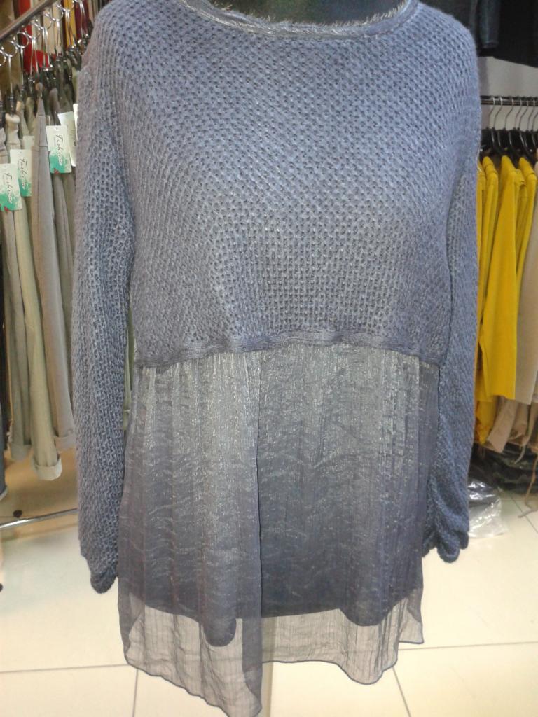 Szaro-niebieska bluzka damska z dzianiny i prześwitującego materiału, wisząca na manekinie. W tle wieszaki z odzieżą w różnych kolorach.