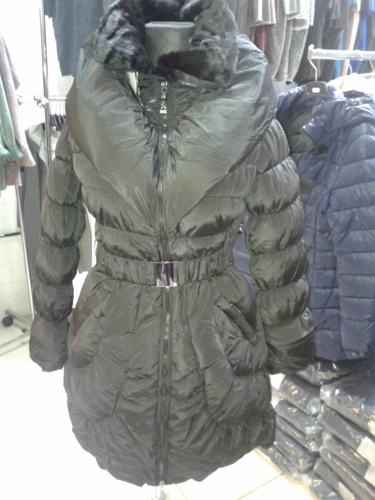 Czarna, pikowana kurtka zimowa z futrzanym kołnierzem i paskiem na manekinie, obok wieszaki z innymi ubraniami w tle sklepu odzieżowego.