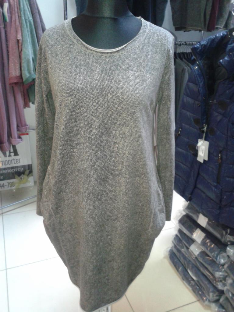 Szara, dzianinowa sukienka tunika z długim rękawem na manekinie, w tle wieszaki z odzieżą i sterta zapakowanych ubrań.