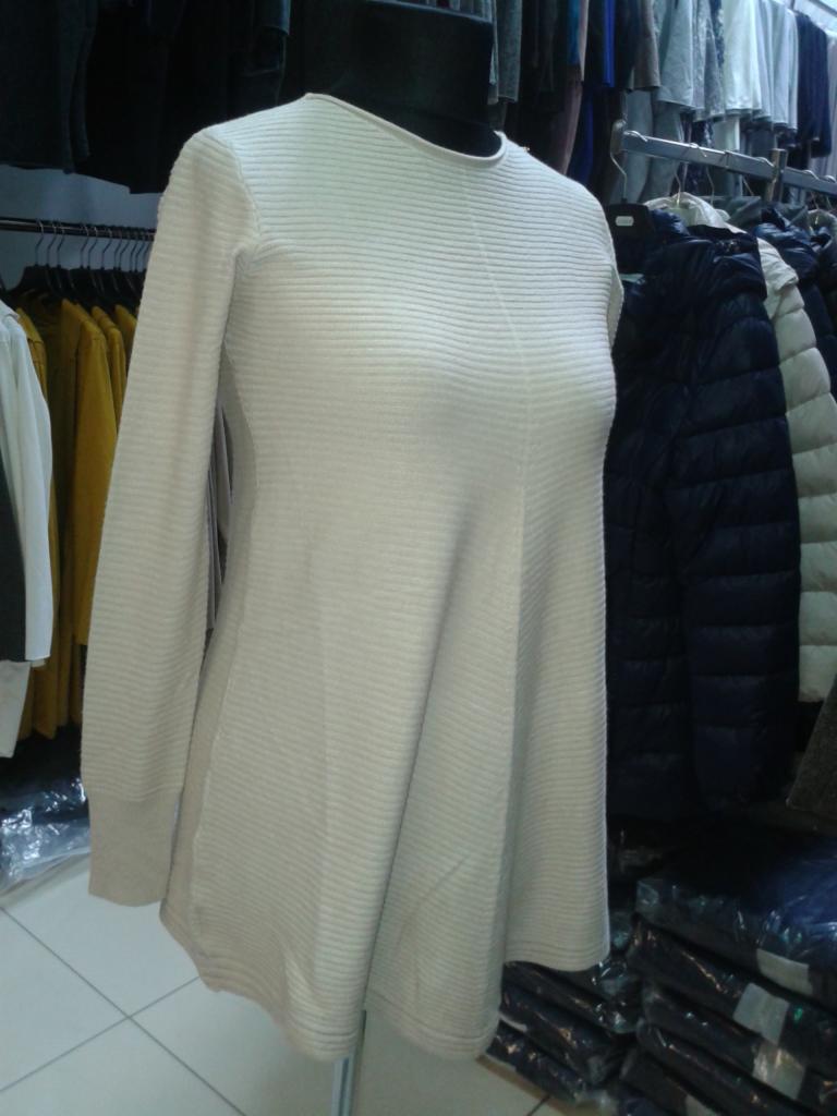 Beżowa tunika z długim rękawem, o prążkowanej fakturze, na manekinie w sklepie odzieżowym. W tle wieszaki z ubraniami i ułożone w stosy paczki z odzieżą.
